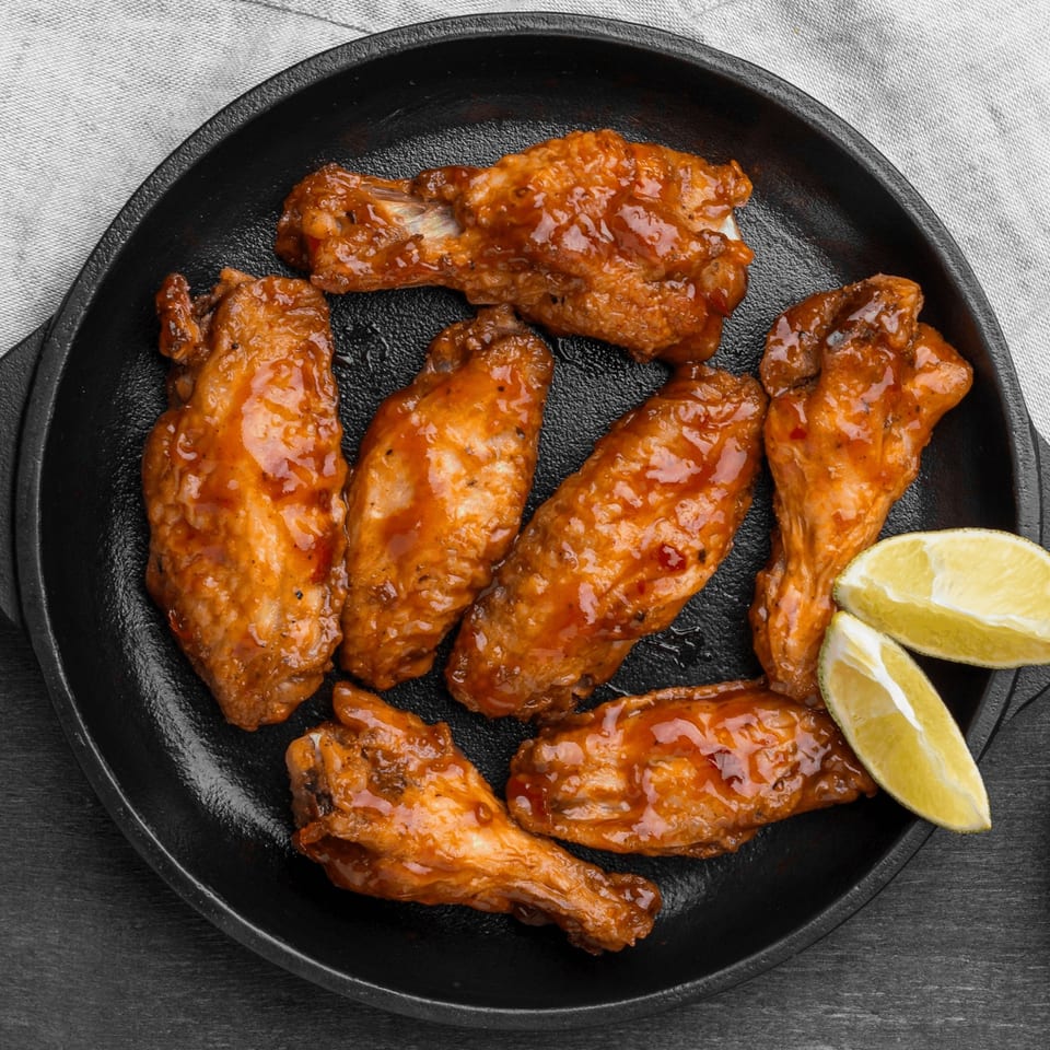 Seven glazed chicken wings with two lime wedges in a black cast iron pan.